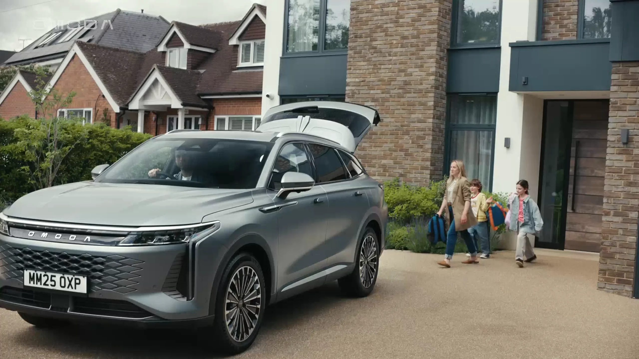 Grey OMODA 9 packed in front of a white and bricked house with a person and two children walking towards the car with another person in the drivers seat with their hand on the wheel